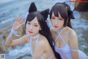 Two women in white bathing suits sitting on a rock by the ocean.